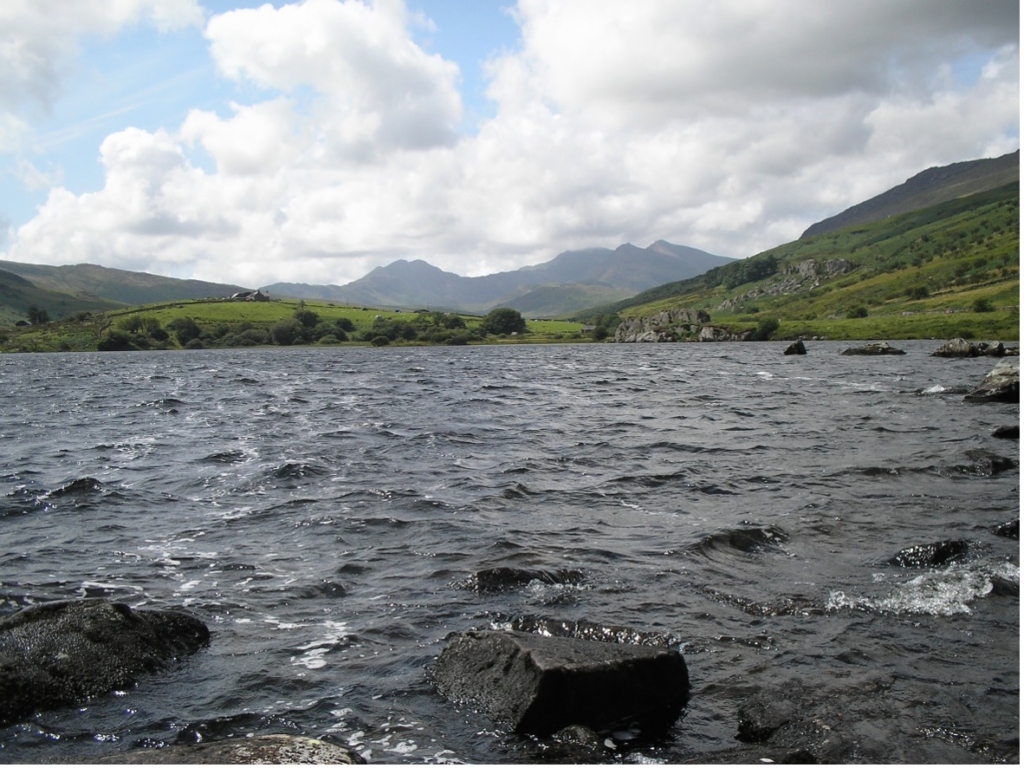 A view of Snowdon across the water to Snowdon, beneath a sky filled with scudding clouds.